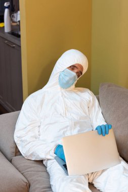 Tıbbi maskeli yorgun bir adam, lateks eldivenler ve dizüstü bilgisayarı kanepede tutuyor. 