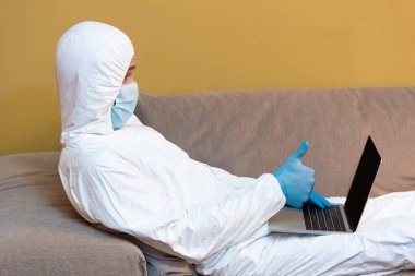 Kanepede dizüstü bilgisayar kullanırken tıbbi maskeli ve tehlikeli madde giysili bir adamın başparmağı görünüyor. 