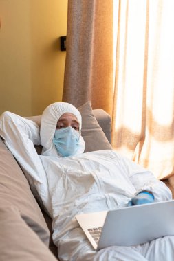 Medikal maskeli ve tehlikeli madde giysili serbest çalışanların evdeki kanepede yatarken dizüstü bilgisayar kullanmaları. 