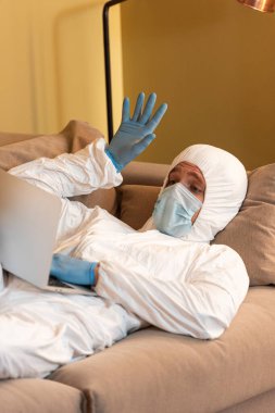 Tehlikeli madde giysisi, tıbbi maske ve lateks eldivenli bir adam evde bilgisayarında görüntülü konuşma yapıyor. 