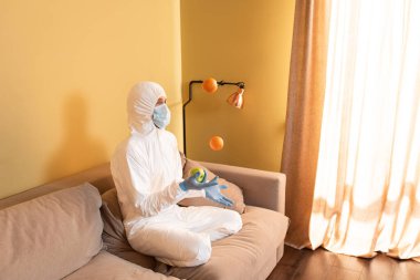 Tehlikeli madde giysisi ve tıbbi maskeli bir adam kanepede meyve hokkabazlığı yapıyor. 
