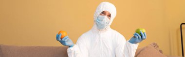 Tehlikeli madde giysisi giymiş düşünceli bir adamın panoramik oryantasyonu ve evde elma ve portakal tutan tıbbi maske. 