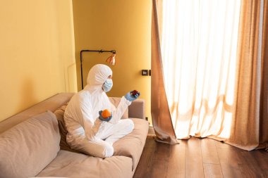 Tıbbi maskeli dalgın bir adam, lateks eldivenler ve tehlikeli madde giysisi koltukta otururken elma ve portakal tutuyor. 
