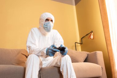 KYIV, UKRAINE - 24 Nisan 2020: Medikal maskeli ve tehlikeli madde giysisi giymiş bir adamın evde bilgisayar oyunu oynaması 
