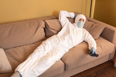 Tıbbi maskeli bir adam, koruyucu giysi ve lateks eldivenler. Evdeki kanepede televizyon seyrediyor. 