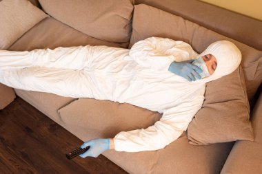 Tehlikeli madde giysisi, tıbbi maske ve lateks eldivenli bir adamın kanepede film izlerken yüksek açılı görüntüsü. 
