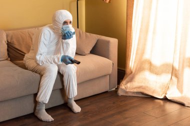 Tıbbi maskeli bir adam, lateks eldivenli ve koruyucu giysili bir adam evdeki koltukta uzaktan kumanda tutuyor. 