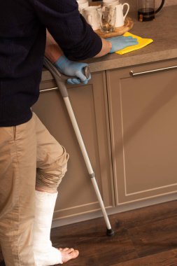 Cropped view of man with plaster bandage on leg cleaning worktop in kitchen  clipart