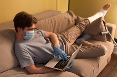 Tıbbi maskeli bir adamın seçici odak noktası ve bacağında alçı bandaj var. Kanepede dizüstü bilgisayar kullanıyor. 