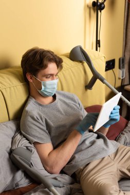 Yatakta koltuk değneklerinin yanında dijital tablet kullanan tıbbi maskeli ve lateks eldivenli engelli bir adam. 