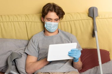 Tıbbi maskeli ve lateks eldivenli bir adam evde koltuk değneklerinin yanında dijital tablet kullanıyor. 