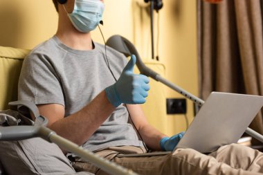 Medikal maskeli ve kulaklıklı adamın seçici odağı başparmağının koltuk değneklerinin yanındaki dizüstü bilgisayarda belirdiğini gösteriyor. 