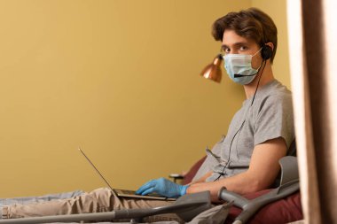 Tıbbi maskeli ve kulaklıklı bir adamın yatak odasındaki koltuk değneklerinin yanında dizüstü bilgisayar kullanırken kameraya bakması.