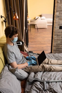 Yataktaki koltuk değneklerinin yanında dizüstü bilgisayar kullanarak tıbbi maske ve kulaklık takan engelli serbest çalışan. 