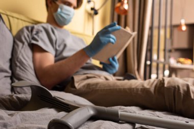 Tıbbi maskeli bir adamın yanında koltuk değneklerinin seçici odağı ve yatakta kitap okuyan lateks eldivenler 
