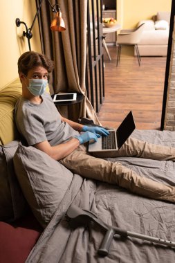 Yatakta koltuk değneklerinin yanında dizüstü bilgisayar kullanan tıbbi maskeli bir adam kameraya bakıyor. 