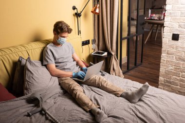 Yatak odasındaki koltuk değneklerinin yanında dizüstü bilgisayar kullanan tıbbi maskeli sakat bir tele-işçi.