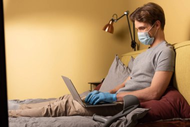 Yatak odasındaki koltuk değneği yanında laptopla çalışan tıbbi maskeli adamın yan görüntüsü.