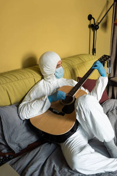 KYIV, UKRAINE - 24 Nisan 2020: Tehlikeli madde giysisi ve tıbbi maskeli adam yatakta otururken akustik gitar çalıyor 