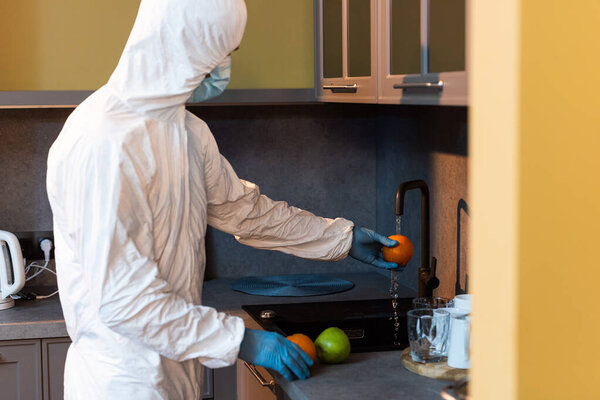Side view of man in hazmat suit and latex gloves washing fruits in kitchen 