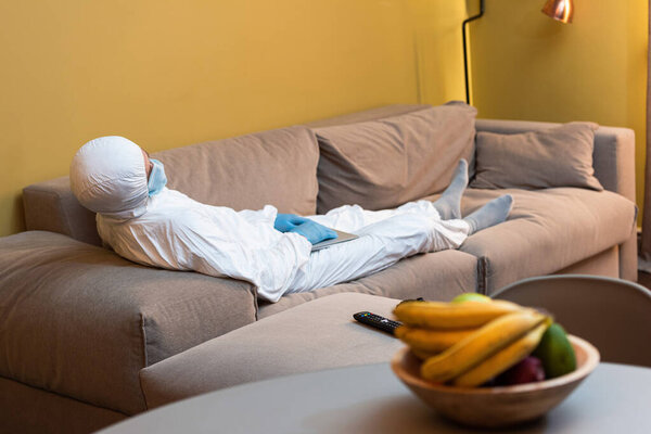 KYIV, UKRAINE - APRIL 24, 2020: Selective focus of man in hazmat suit and medical mask holding laptop near remote controller and gamepad on couch 