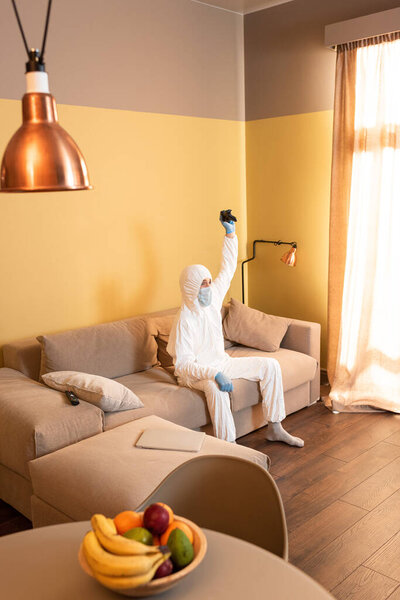 KYIV, UKRAINE - APRIL 24, 2020: Selective focus of man in hazmat suit and medical mask holding joystick on sofa at home 