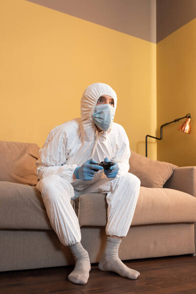 KYIV, UKRAINE - APRIL 24, 2020: Low angle view of man in hazmat suit and medical mask playing video game in living room