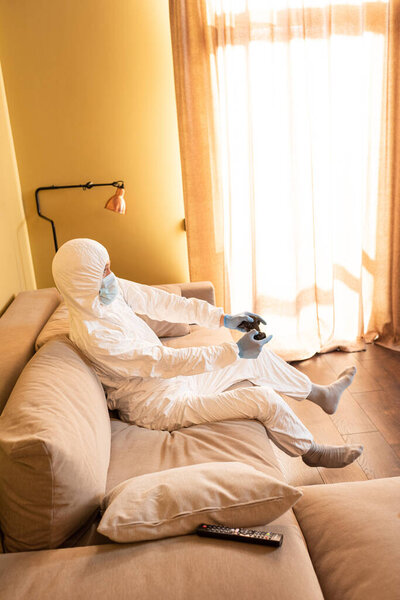 KYIV, UKRAINE - APRIL 24, 2020: Side view of man in hazmat suit and latex gloves playing video game on couch 