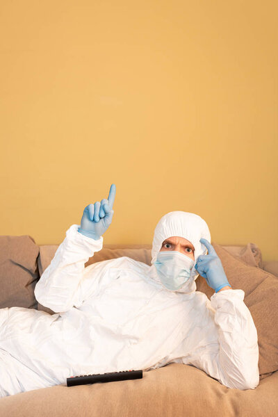 Pensive man in hazmat suit and medical mask pointing with fingers while looking at camera near remote controller on couch 