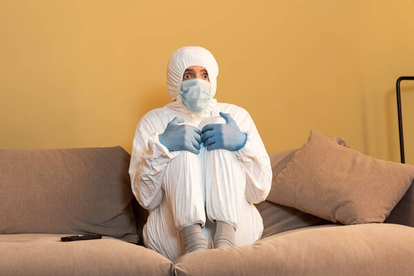 Scared man in hazmat suit and medical mask watching tv on couch 