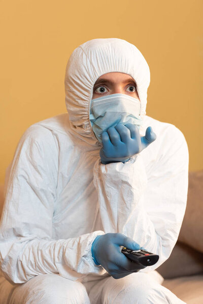 Man in hazmat suit, medical mask and latex gloves holding remote controller on couch 