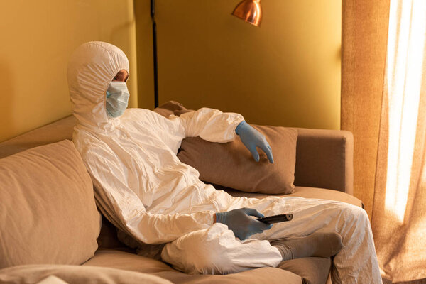 Side view of man in hazmat suit, medical mask and latex gloves holding remote controller on couch 