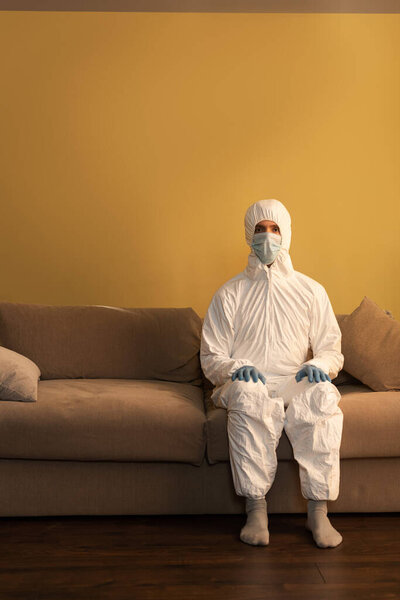 Man in hazmat suit, medical mask and latex gloves looking at camera while sitting on couch 