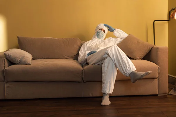 身穿医用面罩 除污服和乳胶手套的男人坐在家里的沙发上 — 图库照片