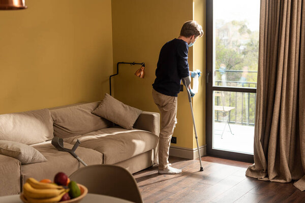 Selective focus of disabled man in medical mask and latex gloves holding detergent near door handle in living room