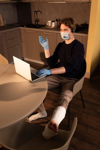 Freelancer in medical mask and plaster bandage on leg pointing with hand while using headset and laptop in kitchen — Stock Image Freelancer Medical Mask Plaster Bandage Leg Pointing Hand While Using Stock Picture