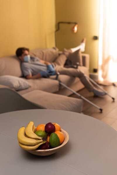 Selective focus of fruits on table and man with broken leg holding remote controller on couch at home 