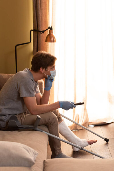 Side view of man in medical mask with broken leg watching movie near crutches on sofa 