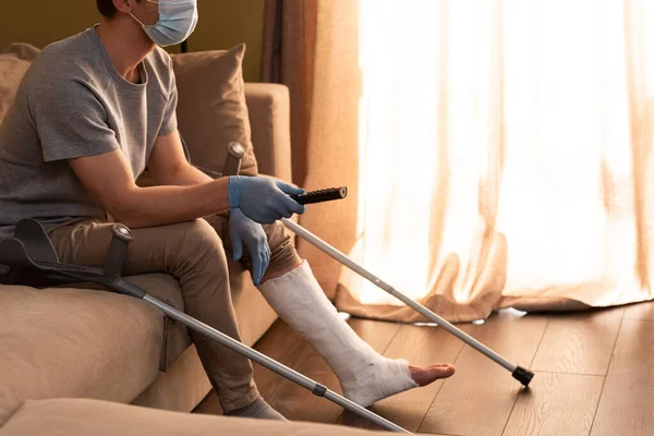 Cropped View Man Medical Mask Broken Leg Clicking Channels Home — Stock Photo, Image