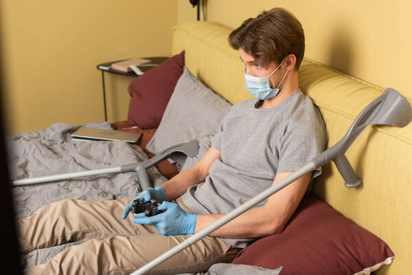 КИЕВ, УКРАИНА - 24 апреля 2020 года: Выборочный фокус инвалида в медицинской маске и латексных перчатках, играющего в видеоигру возле костылей и ноутбука на кровати
 