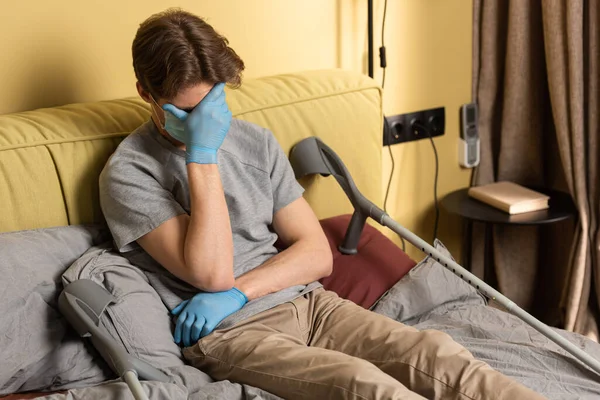 Depressed man in medical mask and latex gloves covering face with hand near crutches on bed