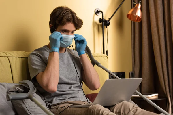 Tıbbi maskeli ve kulaklıklı engelli adam koltuk değneklerinin yanındaki laptopa bakıyor. 