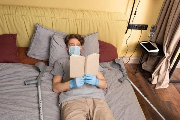 戴着医疗面罩和乳胶手套的残疾人在床上抱着书本时看着相机 — 图库照片