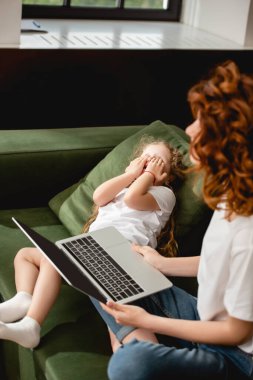 Kıvırcık annenin dizüstü bilgisayarını tuttuğu, duygusal kızının oturma odasında yüzünü örttüğü seçici odak noktası. 