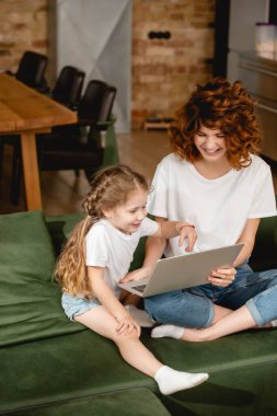 Neşeli kızıl saçlı anne, elinde dizüstü bilgisayarla, mutlu kızının yanında oturma odasında. 