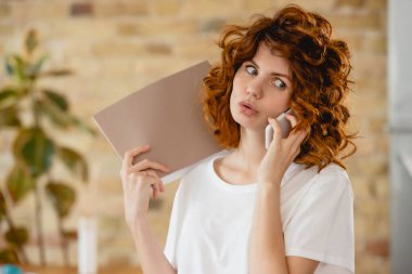 Şaşırmış kızıl saçlı kadın elinde klasör ve akıllı telefondan konuşuyor. 