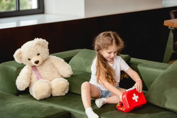 cute child touching toy first aid kit box near teddy bear while playing doctor game