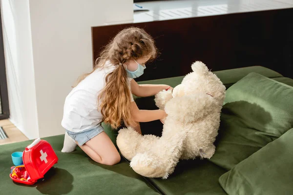戴口罩的可爱小孩儿摸着急救箱旁的泰迪熊 — 图库照片