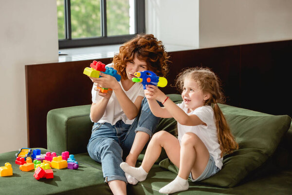 curly mother and daughter with water gun playing in living room 