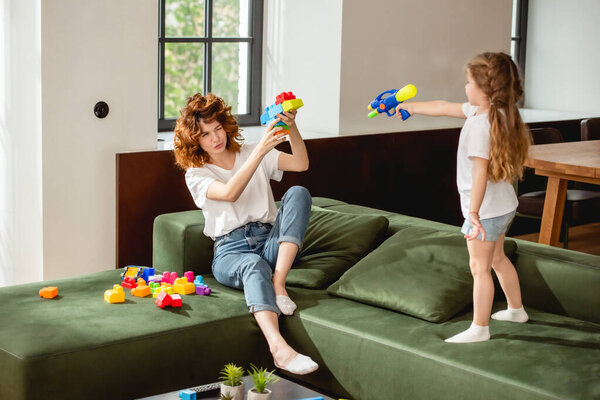 curly mother winking eye and playing with daughter holding water gun in living room 